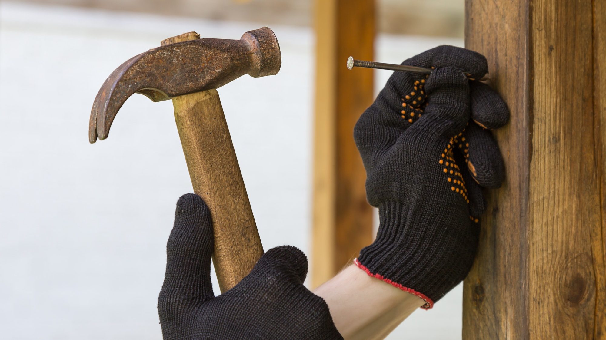 Hand in a construction glove hammers a nail into a wooden beam with a hammer