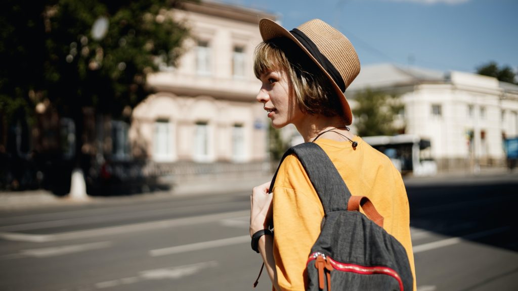 Young girl with headphones in a yellow t-shirt and a straw hat crossing the road with a backpack in